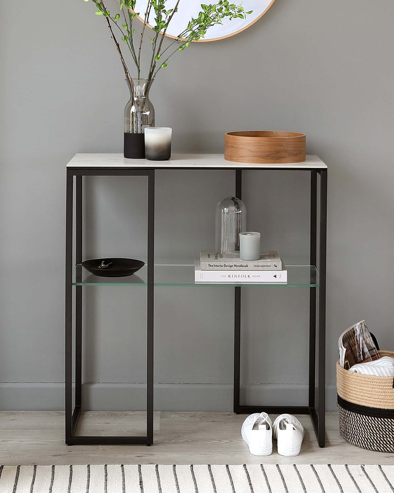 Modern console table with a minimalist design featuring a sleek black metal frame and a light wood tabletop. The table includes one glass shelf mid-way for additional display options. The design is complemented by a woven basket, books, decorative items, and white sneakers placed below and on the table, all set against a muted grey wall with a circular mirror mounted above.