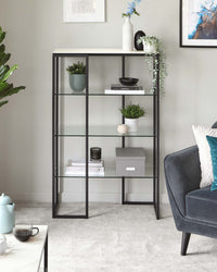 A modern five-tiered shelving unit featuring a sleek black metal frame with rectangular and square shelves of varying heights, complemented by a dark grey upholstered armchair with a textured throw pillow, set against a neutral room decor.