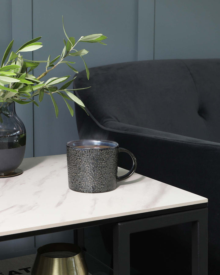 Modern side table with a white marble top and a black metal frame, next to a dark grey upholstered sofa with a smooth, rounded backrest.