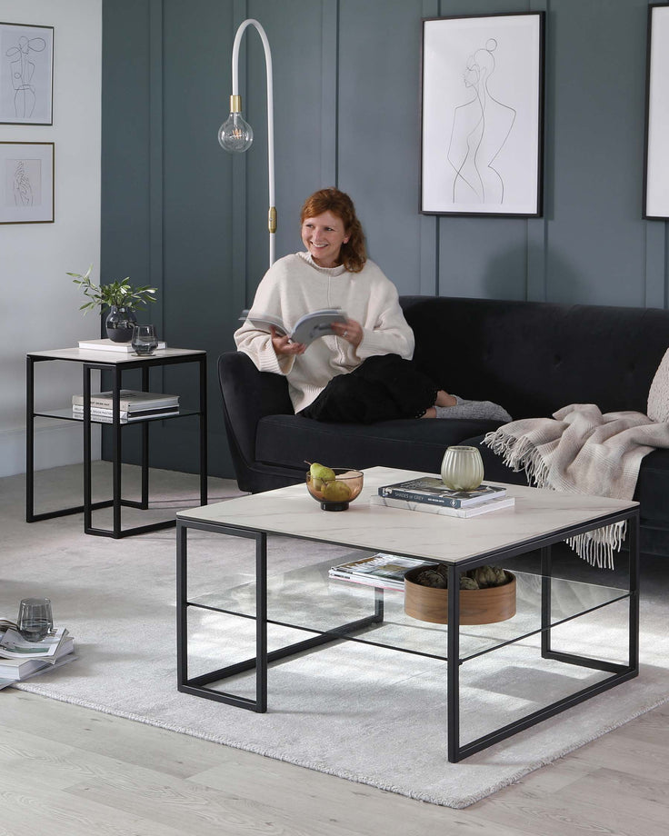 A modern living room setup featuring a sleek black metal-frame sofa with dark upholstery, a rectangular white marble top coffee table with a black metal frame, and a matching black metal-frame side table with a lower shelf displaying books and a plant. The room is decorated with subtle art and soft lighting, emphasizing the contemporary design of the furniture.