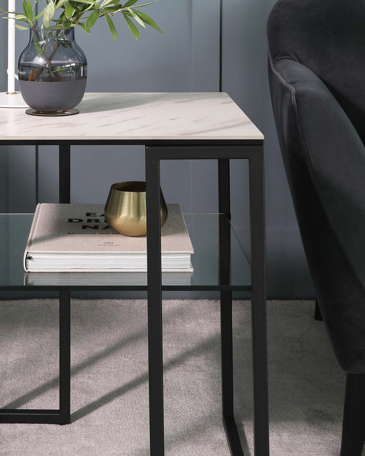 A contemporary marble-top side table with a minimalist black metal frame, accompanied by a partial view of a dark upholstered chair with a curving backrest. The table is styled with a brass decorative bowl and an oversized hardcover book, underneath which lies a segment of a second book or magazine. A clear glass vase with green foliage adds a natural touch to the scene, all resting on a plush light-coloured carpet.