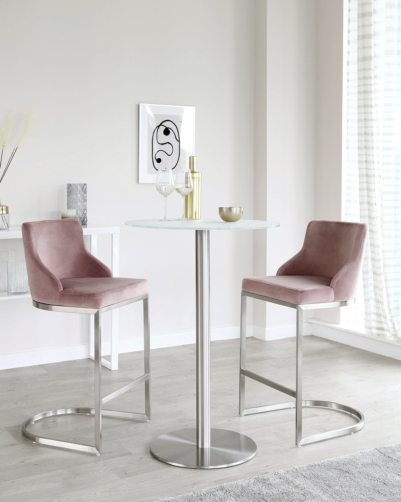 A modern dining set with two mauve velvet upholstered bar chairs featuring a sleek metal frame with a footrest, and a round glass-top table standing on a central polished metal pedestal.