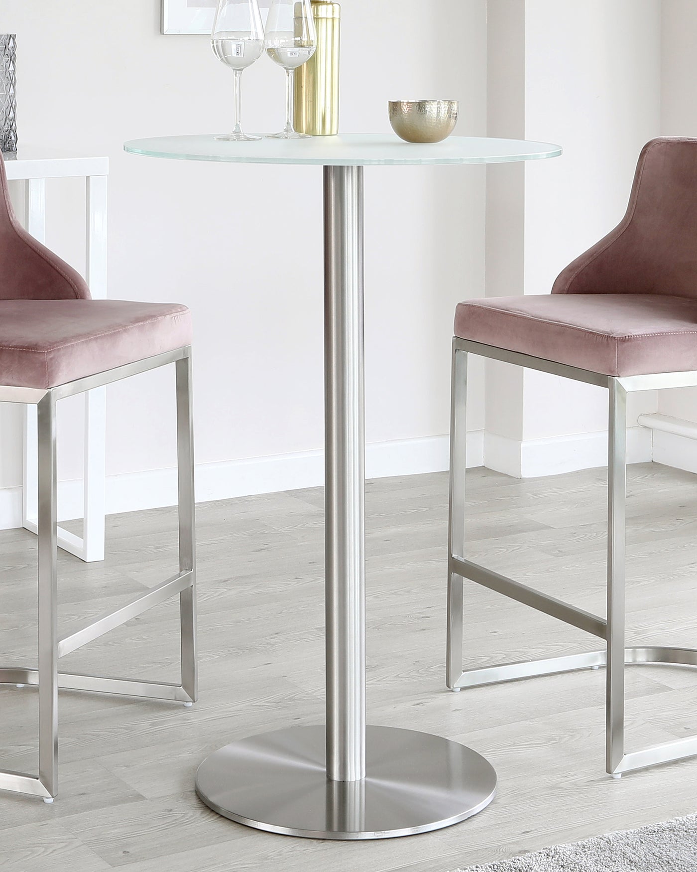 Round glass-top bistro table with a single central metal pedestal and a metal base, accompanied by two modern velvet-upholstered bar stools with metal legs.