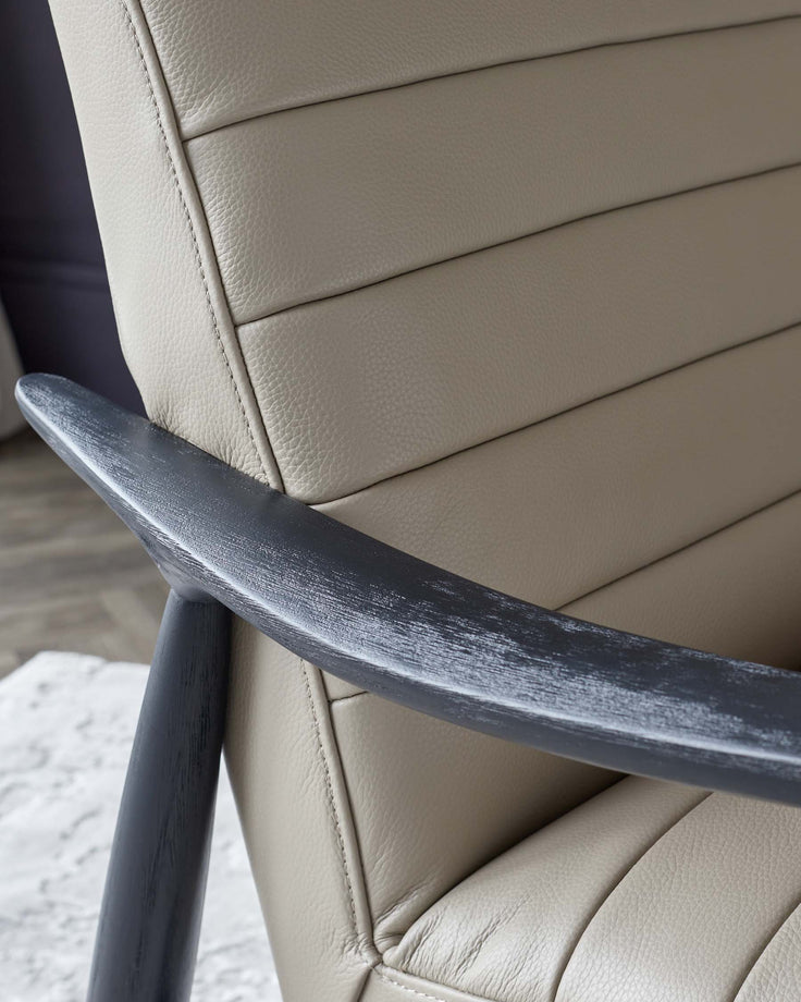 A close-up view of an upholstered chair featuring a cream-colored, textured leather back with horizontal stitching and a dark wooden armrest with a distressed finish.