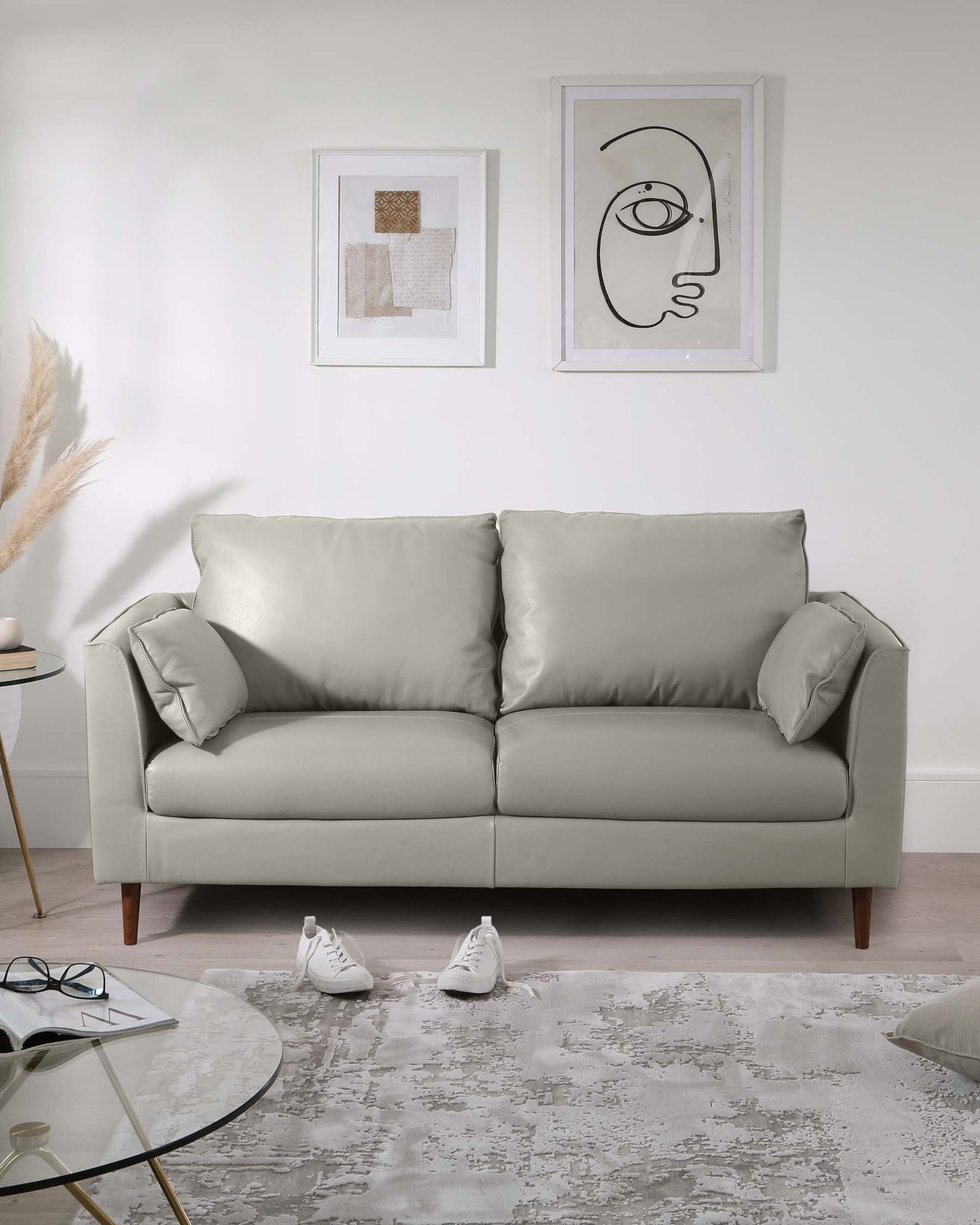 A modern light grey upholstered three-seater sofa with a minimalistic design and plush cushions. The sofa stands on four wooden legs in a mid-century style, positioned on a textured grey area rug near a round glass-top coffee table with a golden rim and black legs. Adjacent to the sofa is a small round side table with a white top and wooden legs displaying a decorative plant. The setting is enhanced by contemporary framed artwork hanging on the wall above the sofa.