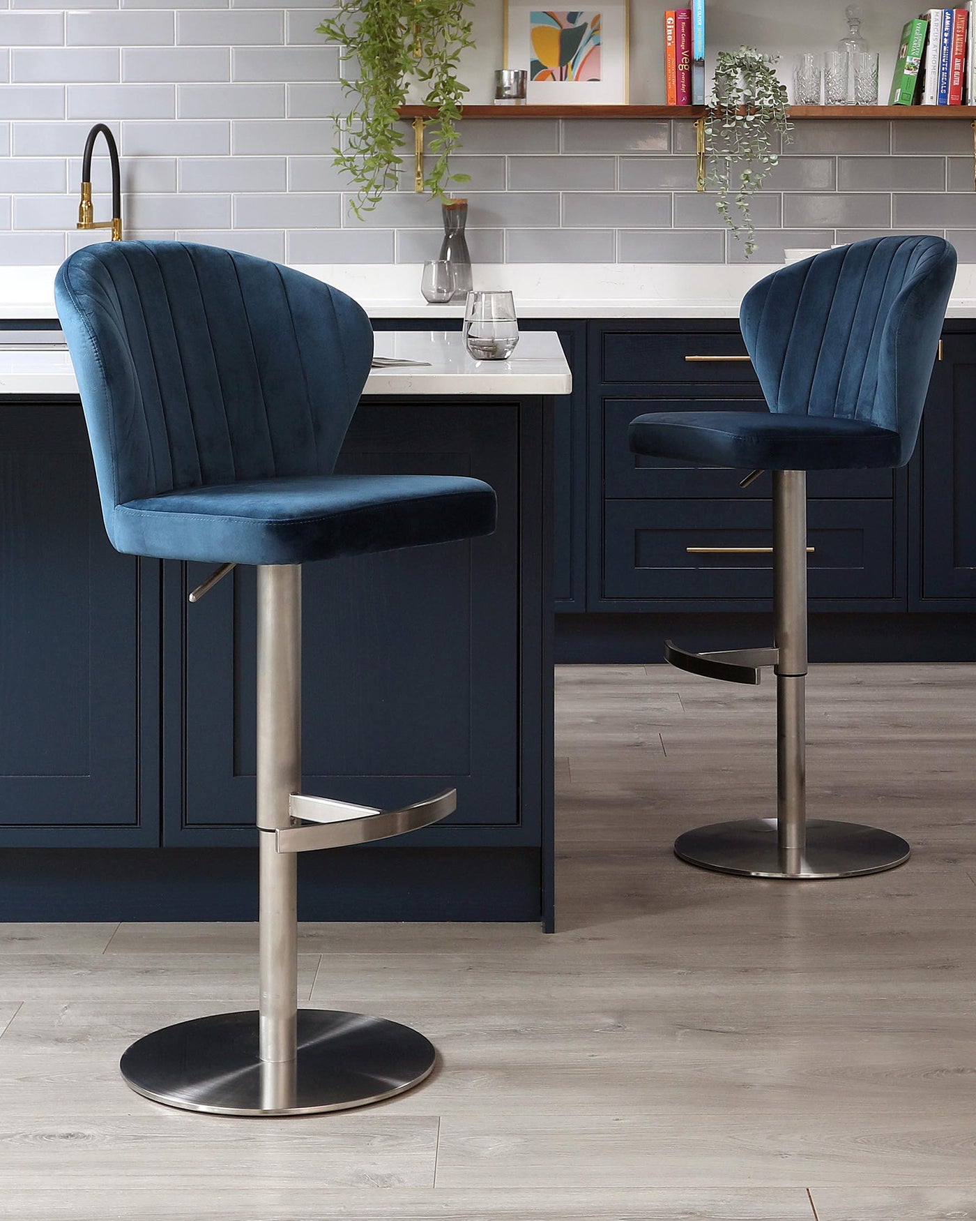 Two elegant contemporary-style bar stools with plush deep blue velvet upholstery and a vertical channel tufted backrest. The stools have an adjustable height mechanism, a circular chrome base, and a built-in footrest for added comfort.
