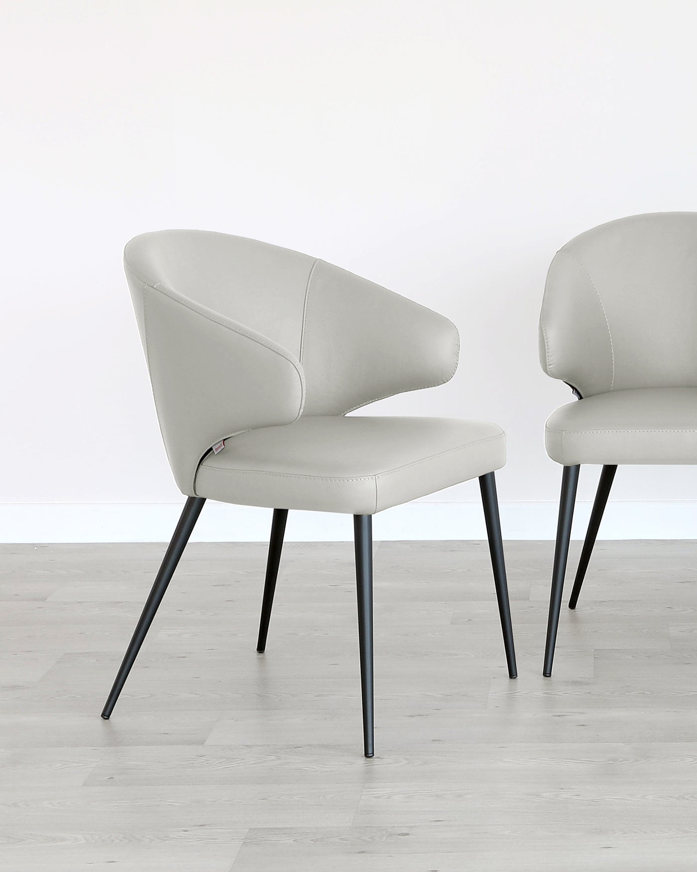 Two modern light grey upholstered dining chairs with curved backrests and angled black metal legs on a light wooden floor against a white backdrop.