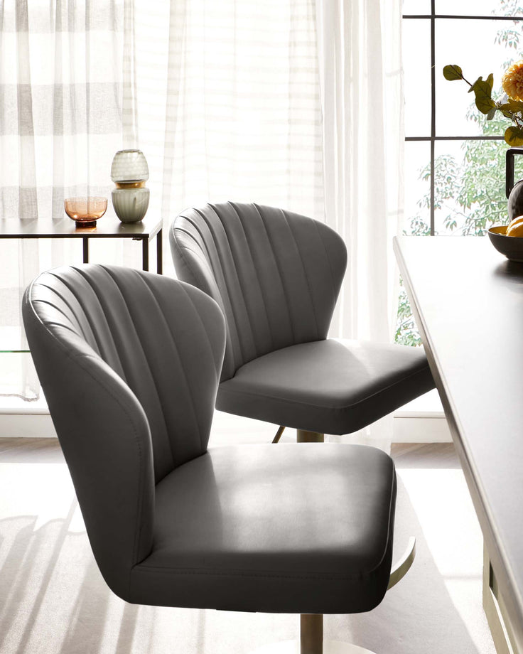Two modern grey upholstered dining chairs with vertical channel tufting and a sleek, black metal base. A small round side table with a black frame and a wooden top, holding decorative vases, is visible in the background. The setting is illuminated by natural light from a large window with sheer curtains.