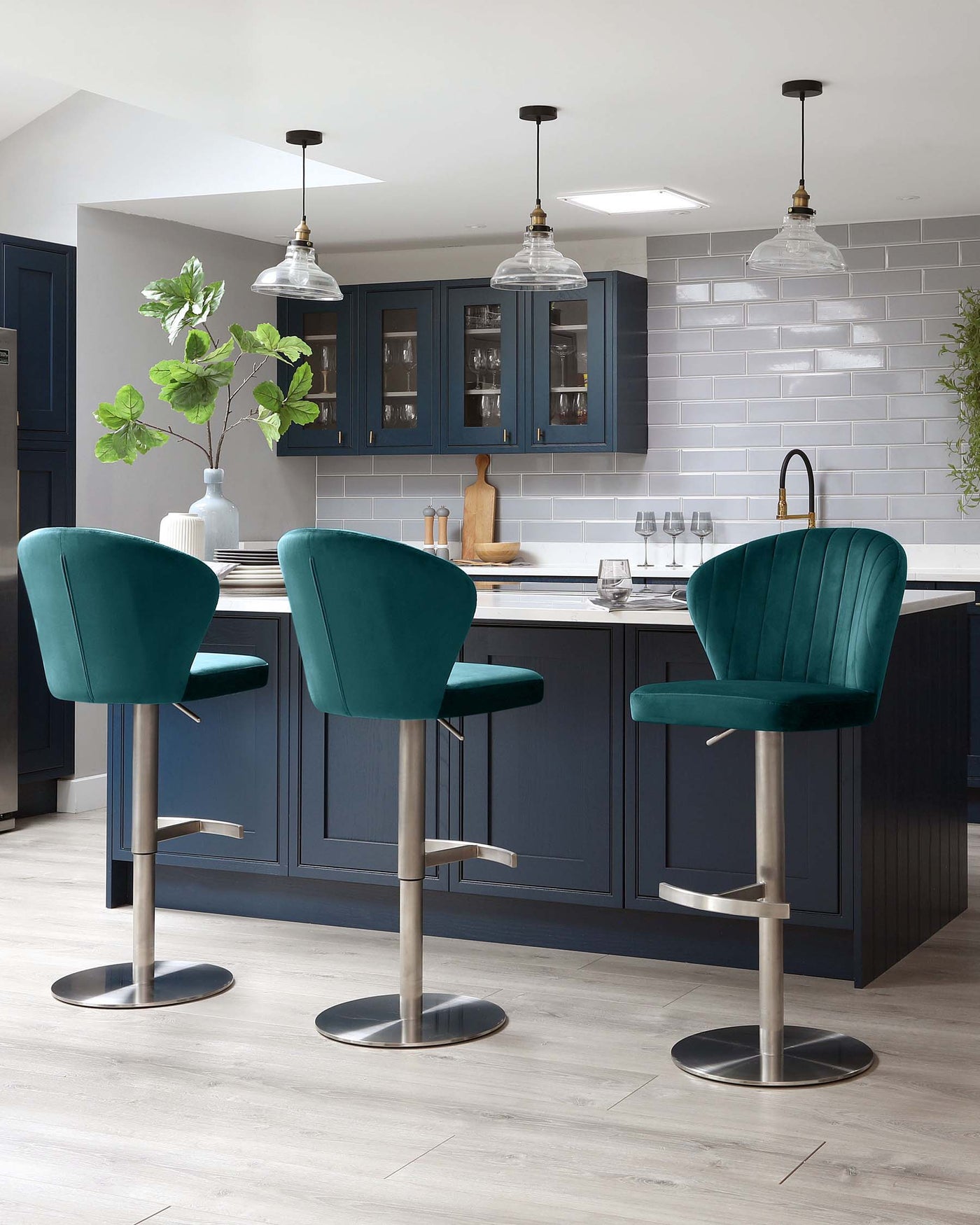 Three modern teal velvet bar stools with low backs and sleek metallic bases positioned around a kitchen island with navy cabinetry and white countertops.