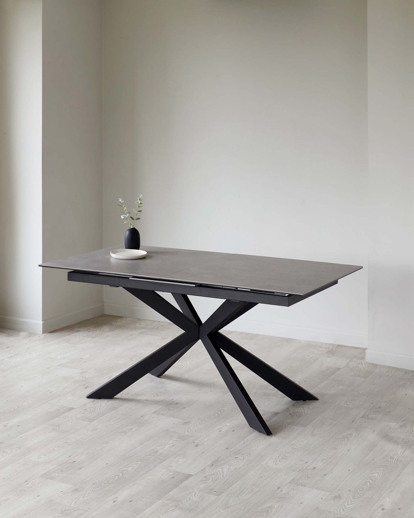 Modern minimalist rectangular dining table with a matte black tabletop and a sculptural black metal base, displayed in a room with light grey walls and pale wooden flooring. A small black vase with a sprig of greenery is placed on the table.