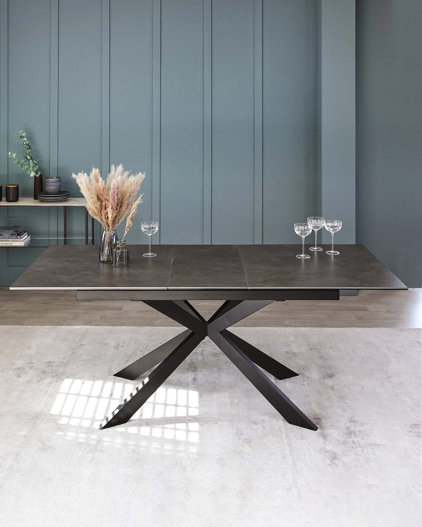 Modern minimalist dining table with a dark, stone-like tabletop and a striking X-shaped metallic base, contrasted against a muted teal wall and light grey floor.