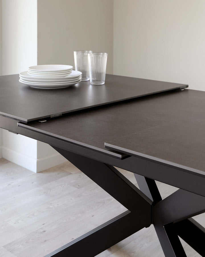 A modern, minimalist dining table with a matte black finish, featuring a sleek tabletop and bold, angular legs crossing asymmetrically for a contemporary look. The table is accessorized with a stack of white plates and clear glasses to highlight its elegance and usability.