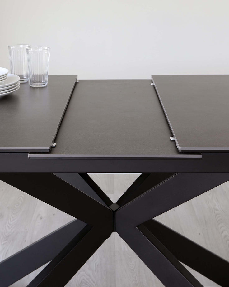 Modern black extendable dining table with a matte finish tabletop and a distinctive X-shaped metallic base, paired with white porcelain dishes and clear drinking glasses on top.