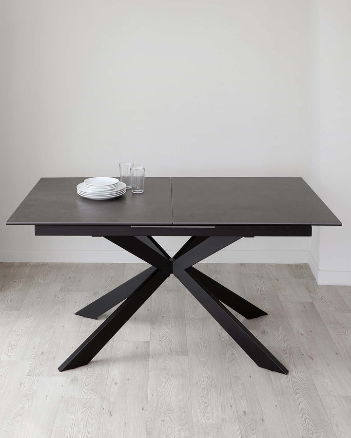 Modern expandable dining table with a matte black finish and a unique, sculptural cross leg design. The table is set against a neutral backdrop, accentuating its clean lines and contemporary aesthetic.