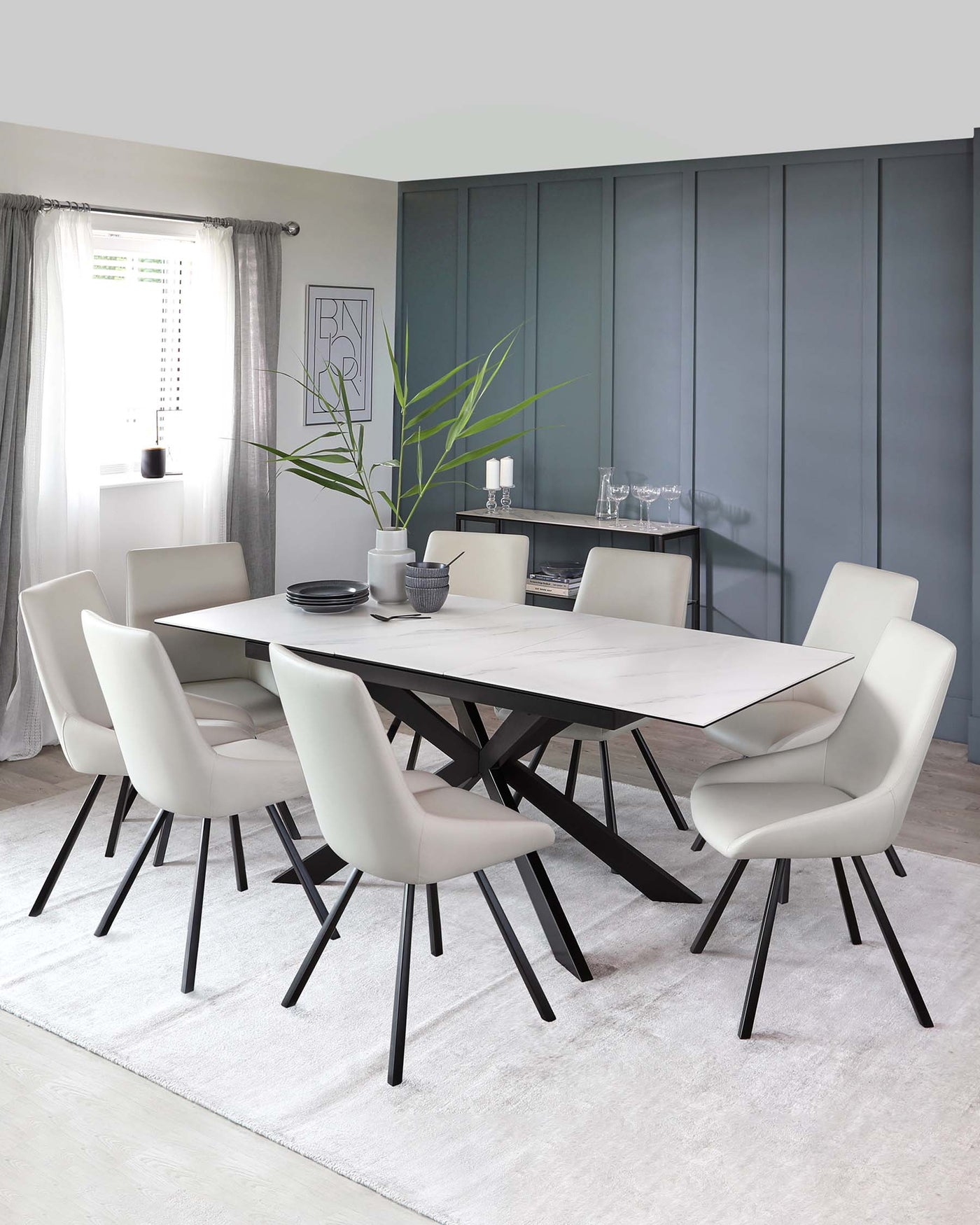 Modern dining room set featuring an oval white marble table with a unique angular black base and six curved, cream-upholstered chairs with slim black legs, arranged on a plain light grey area rug. A sleek black sideboard is in the background.