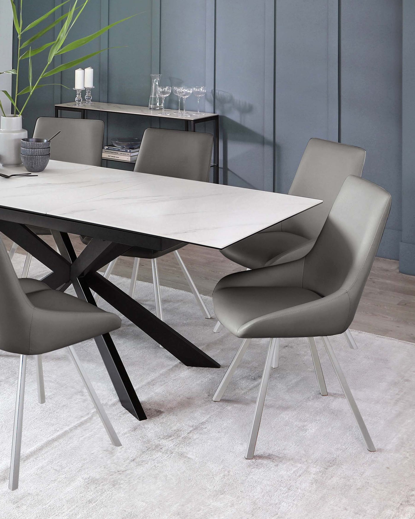 Modern dining room furniture featuring a rectangular table with a white marble top and an angular black base, paired with four sleek grey upholstered chairs with metal legs. A minimalist style sideboard with a dark finish is in the background, accessorized with simple decor.