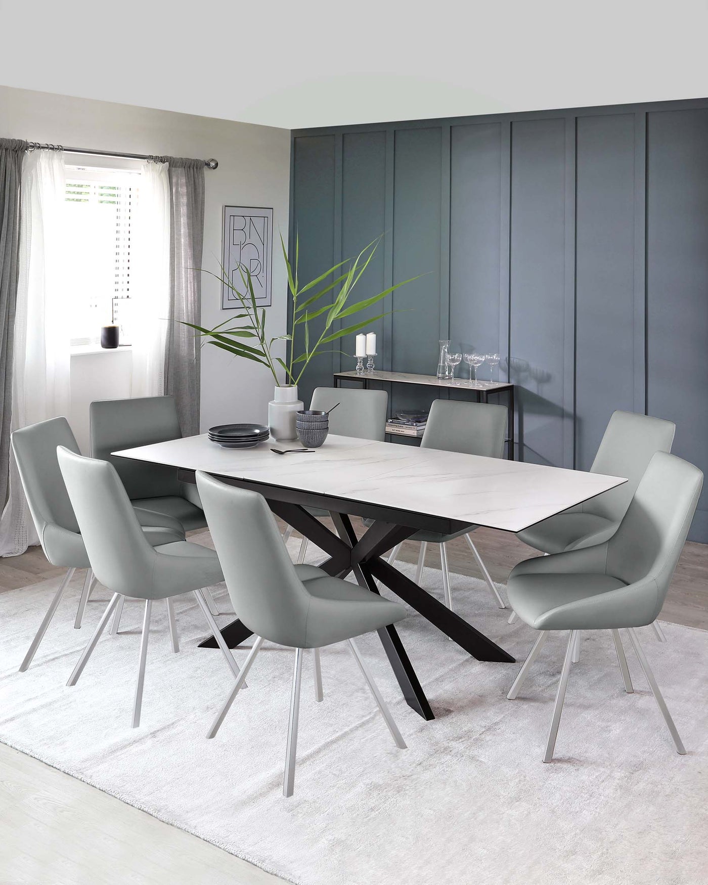 Modern dining room set featuring a rectangular table with a light-coloured tabletop and a striking dark angular base. Surrounding the table are six contemporary-style chairs with sleek light grey upholstery and slender metal legs. The set is displayed on a soft white area rug.