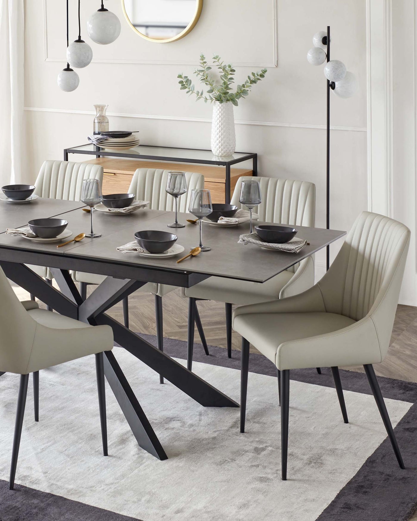 Modern dining room set including a rectangular table with a dark grey surface and angular black metal legs, accompanied by six cream upholstered chairs with vertical stitching and black tapered legs. A sleek black sideboard with a wooden front occupies the background, complemented by a round mirror with a golden frame on the wall above it. The set is arranged on a patterned area rug with grey tones.