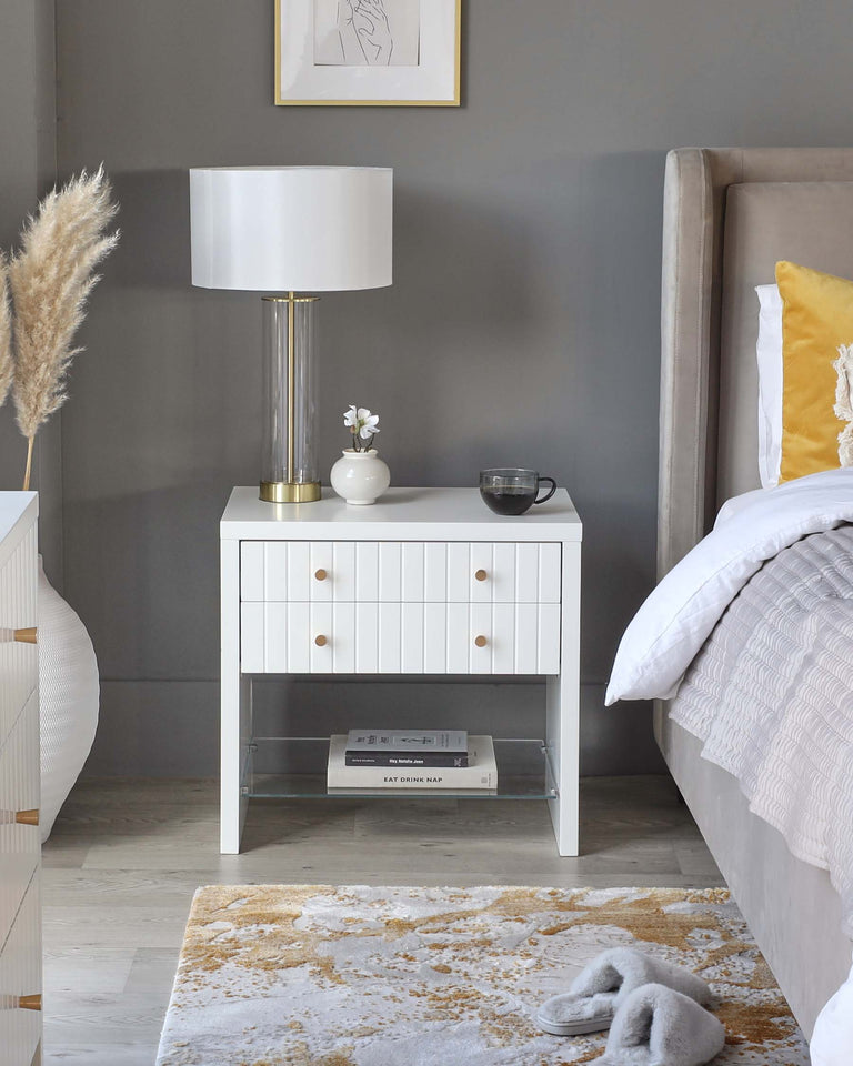 A stylish contemporary bedside table with a clean white finish, featuring three drawers with round, gold-coloured knobs. An open lower shelf offers additional space for books or decor items. The table is complemented by a luxurious upholstered bed with a taupe headboard, partially visible on the right. Both pieces rest on a textured cream and gold area rug, creating a warm and inviting bedroom atmosphere.