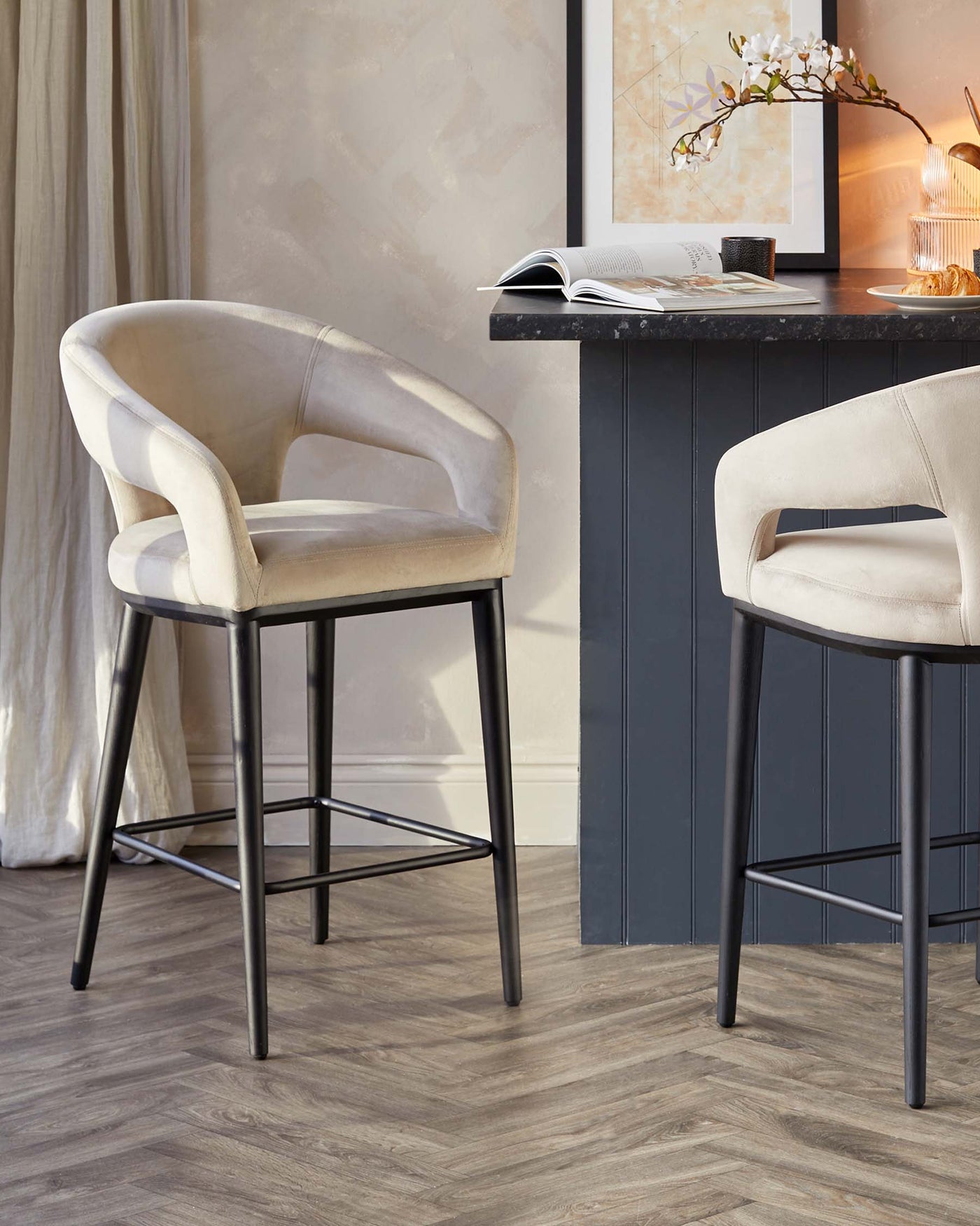 Elegant contemporary bar stools with plush beige velvet upholstery and a curved backrest, featuring sleek black metal legs with a geometric footrest design.
