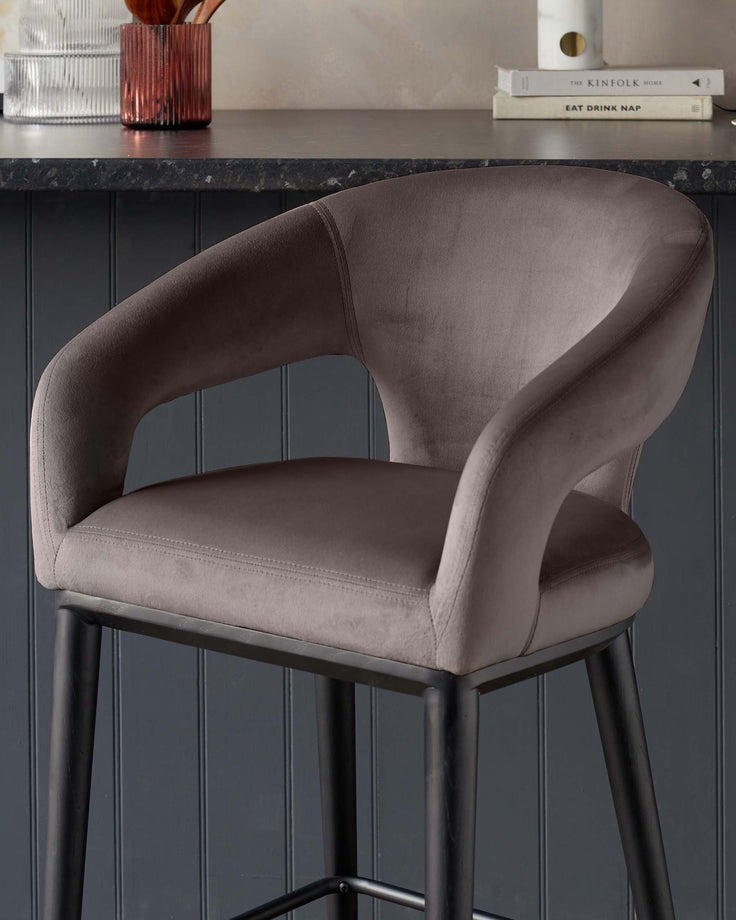 Elegant modern barstool with a curved backrest and cushioned seat upholstered in a smooth, taupe velvet fabric, supported by slender black metal legs with a simple footrest.