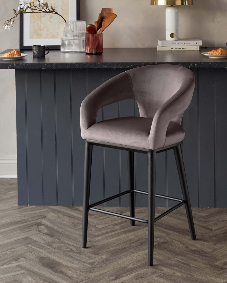 Elegant modern bar stool with a curved, velvet-upholstered backrest and seat in a soft grey tone, featuring sleek black metal legs with a footrest, set against a kitchen counter backdrop.