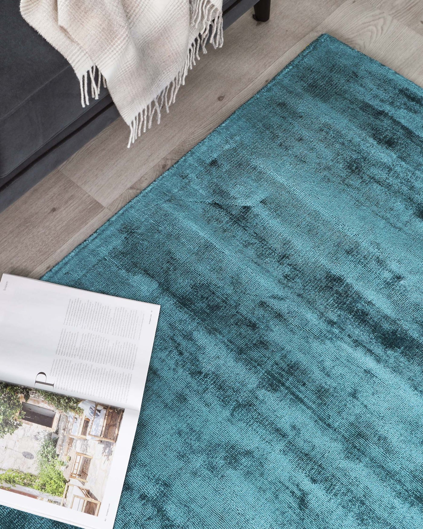 A textured turquoise area rug with a gradient effect, paired with a partial view of a modern sofa's leg covered with a light beige throw blanket with fringes. An open magazine rests on the rug.