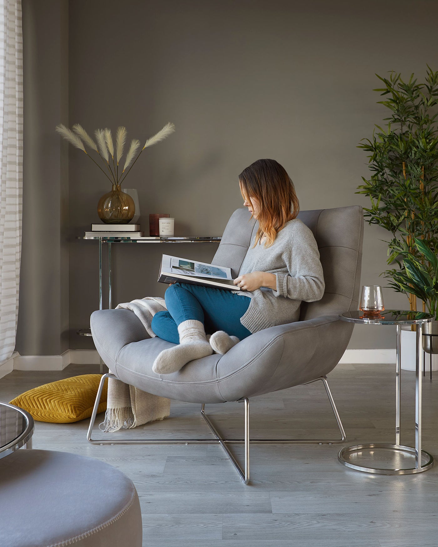Modern room featuring a light grey upholstered lounge chair with a high back, cushioned seat, and sleek metal legs. Next to it, a round glass side table with a polished metallic frame. A cylindrical grey fabric ottoman is also visible in the foreground. The scene conveys a contemporary, cosy atmosphere ideal for reading and relaxation.