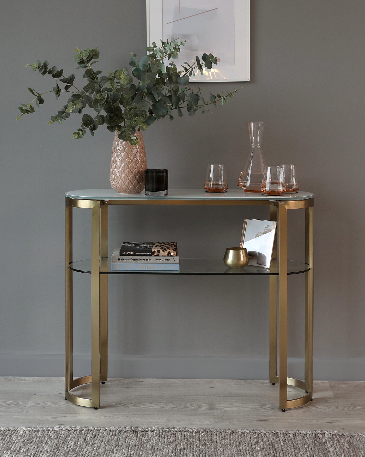 Elegant console table with a sleek, gold-finish metal frame and two tiers featuring glass surfaces, set against a grey wall on a light wooden floor.