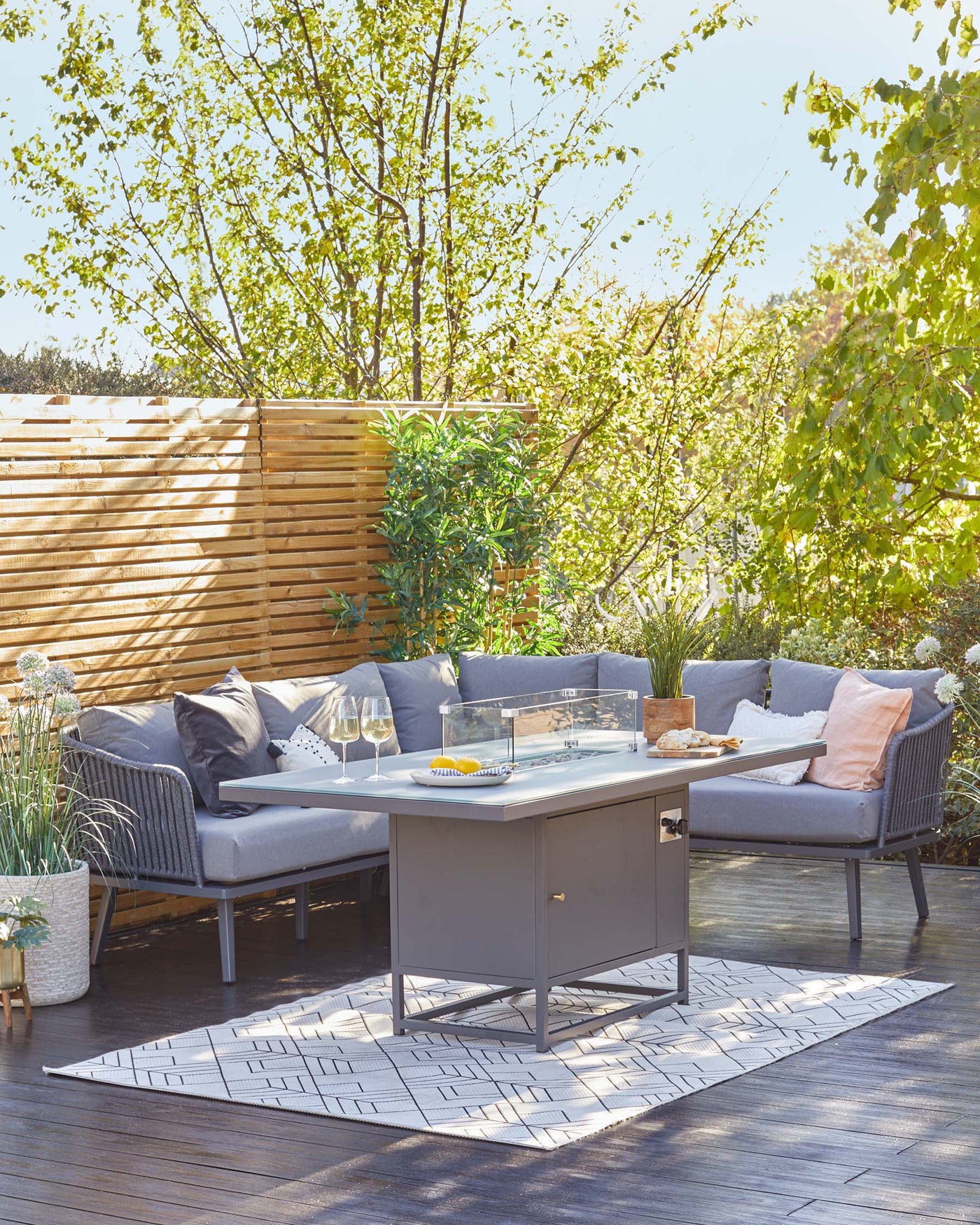 Outdoor patio set featuring a modern, grey sectional sofa with comfortable cushions and pillows, a sleek rectangular coffee table with a glass top and built-in storage, paired with a geometric-patterned rug. The setting is complemented by greenery, adding a natural and serene backdrop.