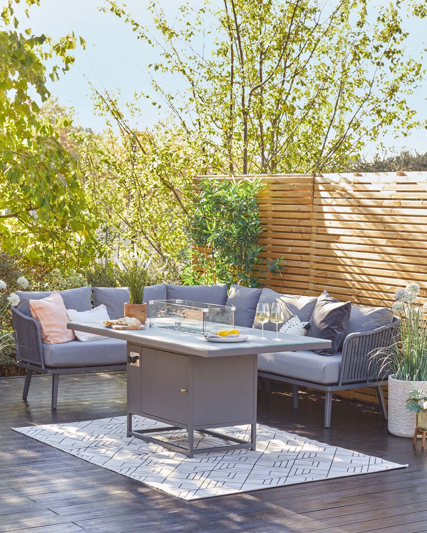 Outdoor patio furniture set featuring a modern grey sectional sofa with plush cushions, complemented by a sleek grey rectangular metal coffee table with a clear glass top. The set rests on a dark wooden deck, accented by a geometric-patterned outdoor rug beneath the table.