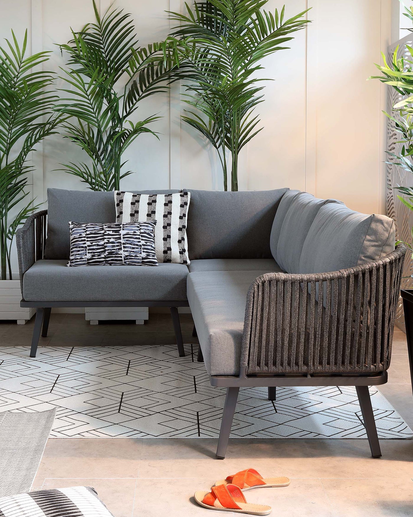Modern charcoal grey sofa with clean lines and a textured weave, featuring dark wooden legs, complemented by an assortment of pillows in geometric and striped patterns. Partial view of a matching chair with a vertical rope back design and similar dark legs to the side of the sofa. The furniture is positioned on an area rug with a black and white geometric pattern, adding to the contemporary aesthetic. Decorative green potted plants create a natural backdrop.