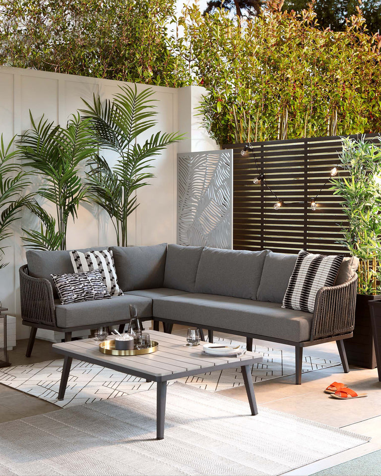 Outdoor furniture set including a grey sectional sofa with white and black patterned accent pillows, and a coordinating grey rectangular coffee table. The set features a modern design with clean lines and is arranged on a neutral outdoor rug, accented with potted green plants.