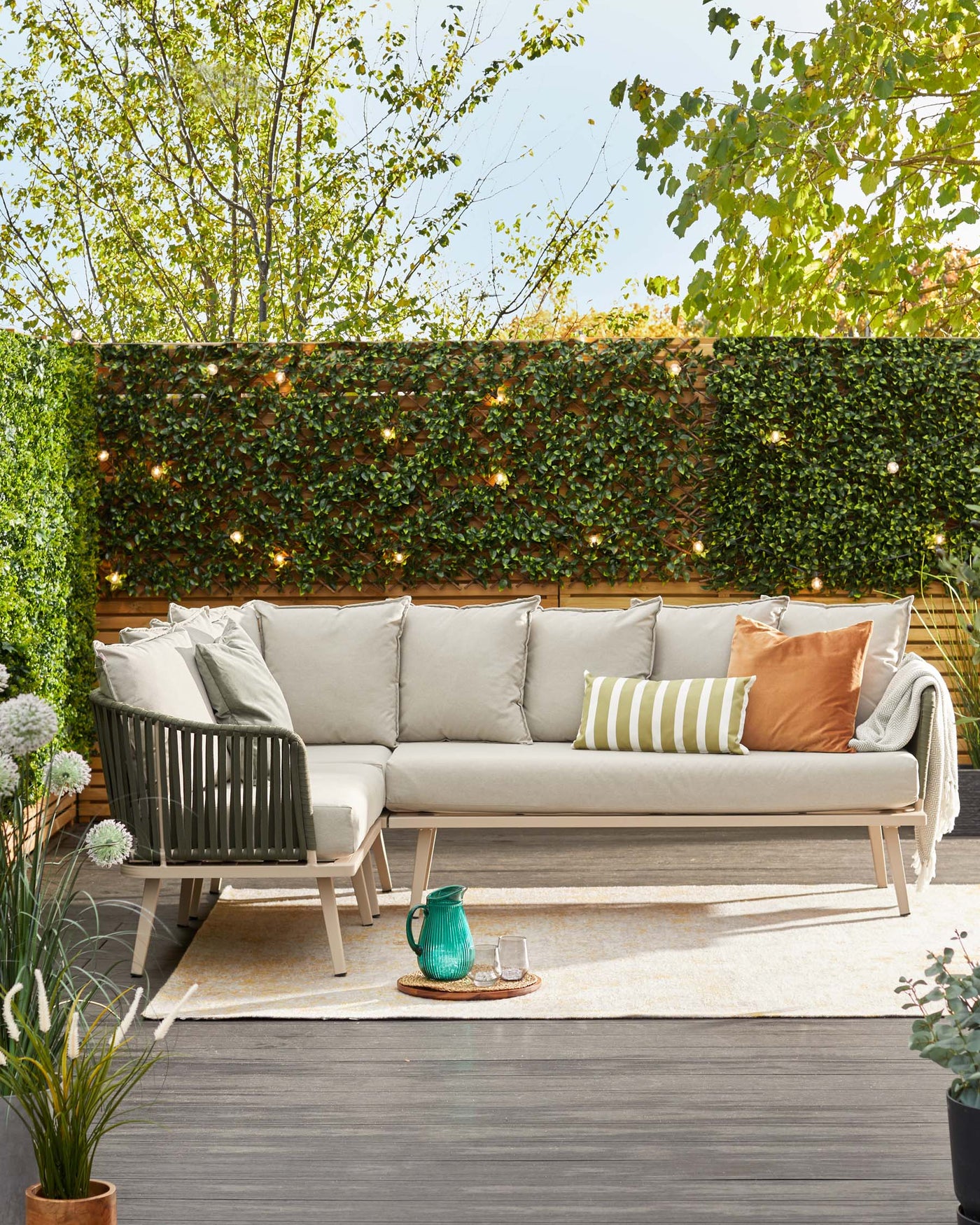 Outdoor corner sofa with light beige cushions on a beige minimalist frame, accompanied by an assortment of throw pillows in beige, green stripes, and copper tones. A textured beige area rug under the sofa, with a glass pitcher and glasses set on a small round tray placed on the rug.