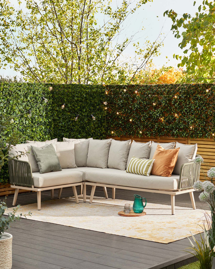 Outdoor sectional sofa with a modular design, featuring a light wooden frame and plush, cream-colored cushions, complemented by additional green and tan throw pillows. The set includes an integrated corner table with vertical slats. The arrangement is positioned on a patterned beige outdoor rug, with a small rattan tray and a green glass pitcher set atop it.