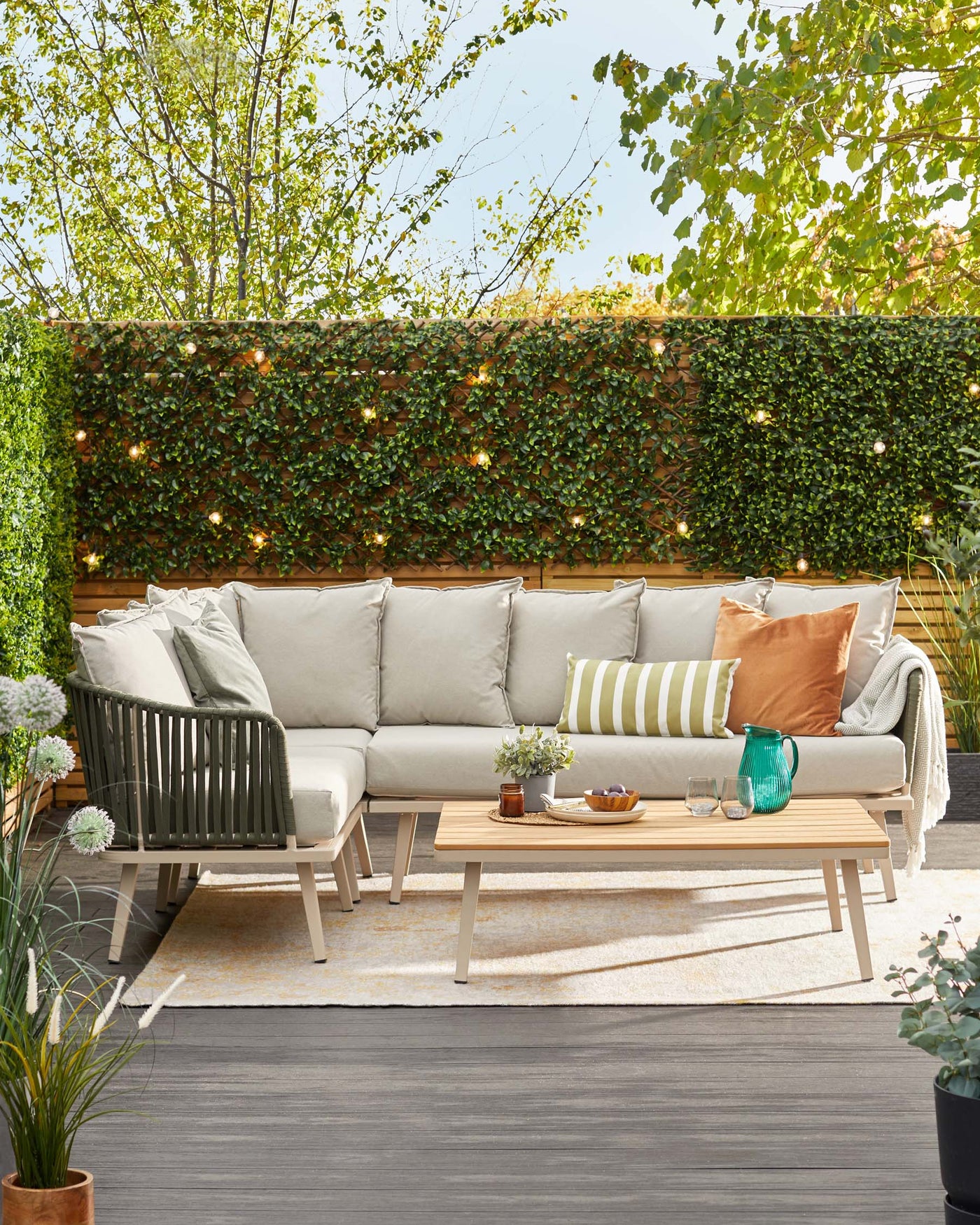 Outdoor furniture set featuring a modern beige upholstered sectional sofa with light grey and accent pillows, a matching wooden armchair with slatted design, and a low-profile rectangular wooden coffee table. The set is arranged on an outdoor rug in a cosy patio setting surrounded by lush greenery and hanging string lights.