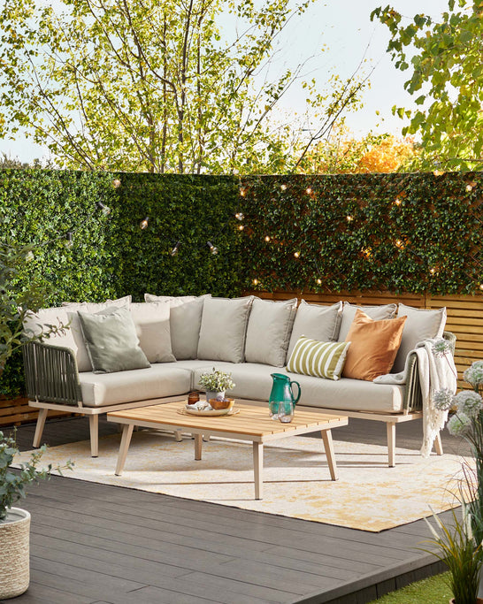 Outdoor corner sectional sofa with plush, neutral-tone cushions and a selection of decorative pillows accompanying a matching wooden rectangular centre table on a decked patio with outdoor rug, amidst lush greenery.
