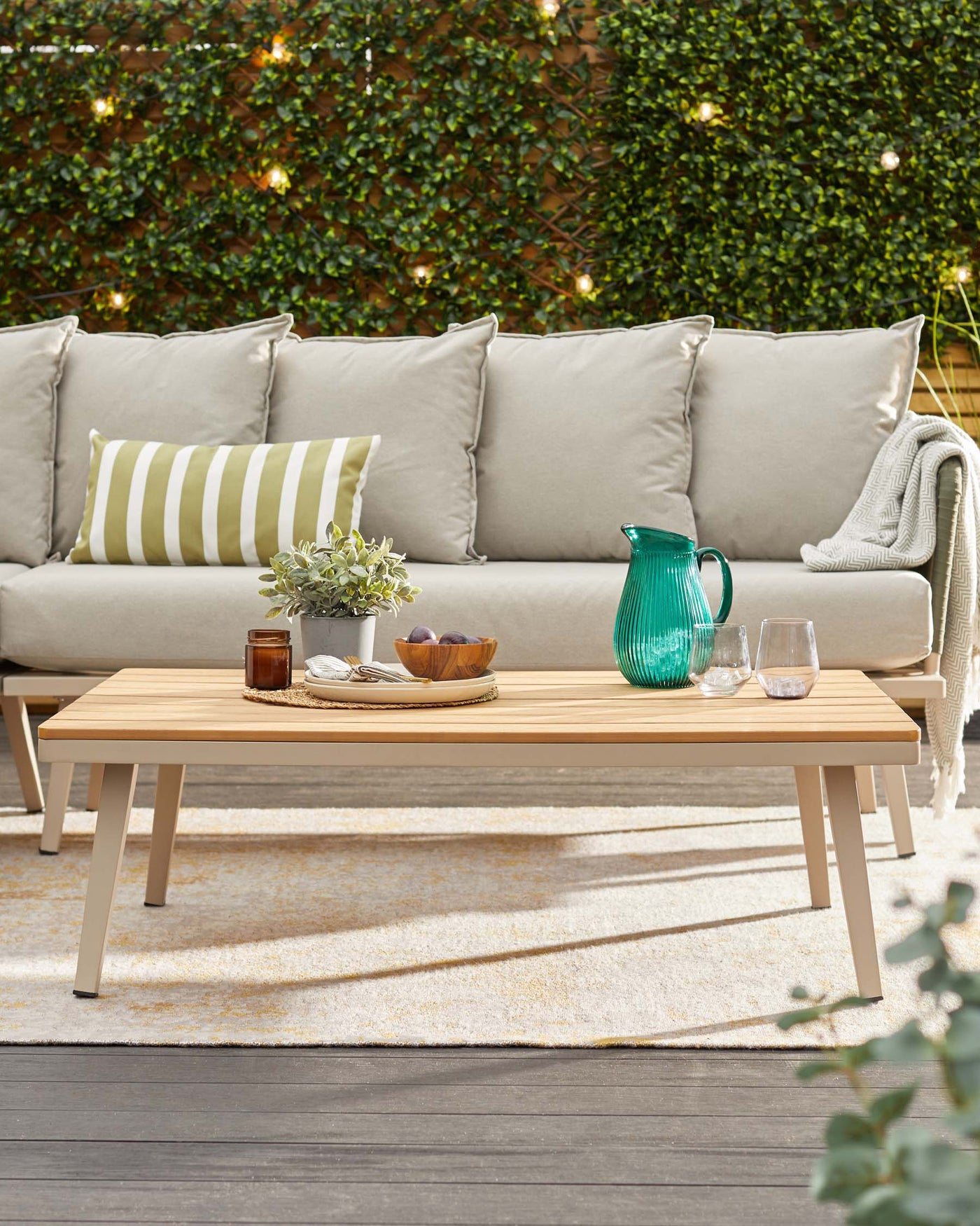 Outdoor furniture set featuring a modern beige sofa with plush cushions and a light grey wooden coffee table on a cosy patio area. The sofa is accented with a mix of solid and striped throw pillows, and a knit blanket for additional comfort. The coffee table is adorned with a plant, a teal glass pitcher, and a wooden bowl with fruits, creating a welcoming entertainment space.