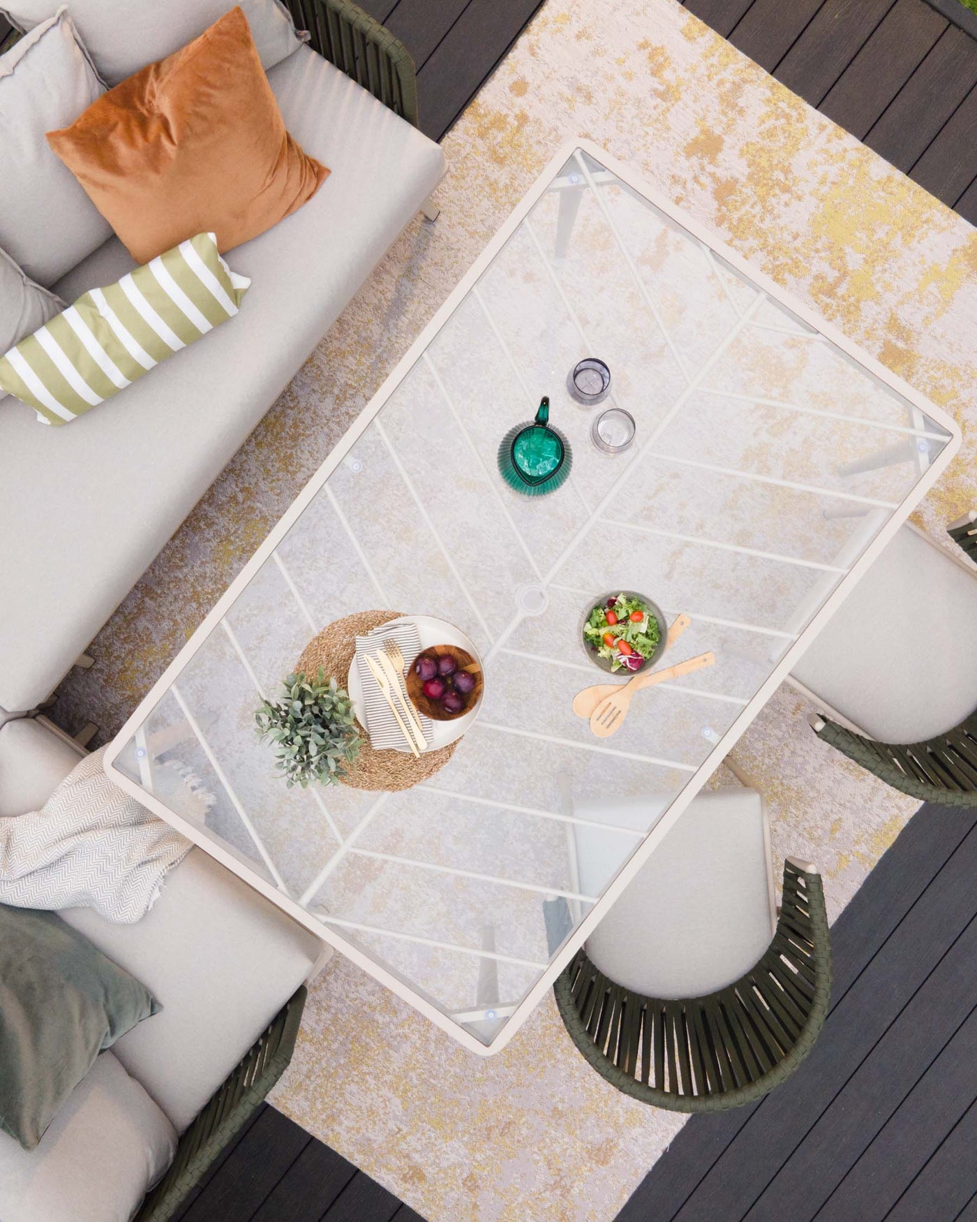 Top-down view of a modern outdoor patio furniture set including a minimalist white sofa with grey cushions and assorted decorative pillows, accompanied by a geometric white metal coffee table with a clear glass top. The set is complemented by two unique olive-green accent chairs with vertical strap design and matching seat cushions. The ensemble is arranged on an elegant mustard and cream area rug, creating an inviting and stylish outdoor living space.