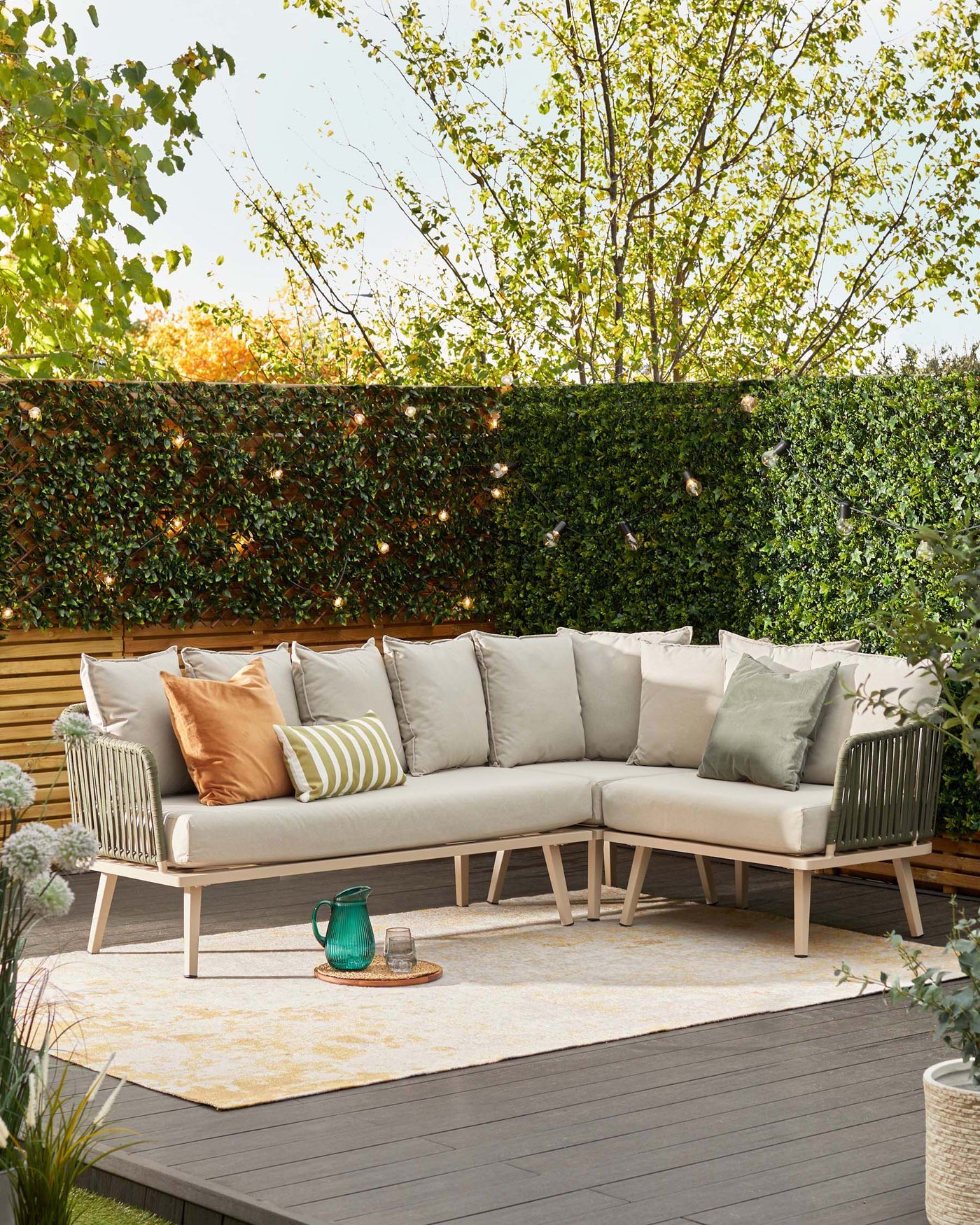 Contemporary outdoor sectional sofa with beige cushions and accent pillows in various shades of green and beige, positioned on a patterned cream rug. A blue-green glass lantern and small glass are set on a round rattan tray, placed on the wooden deck next to the sofa.