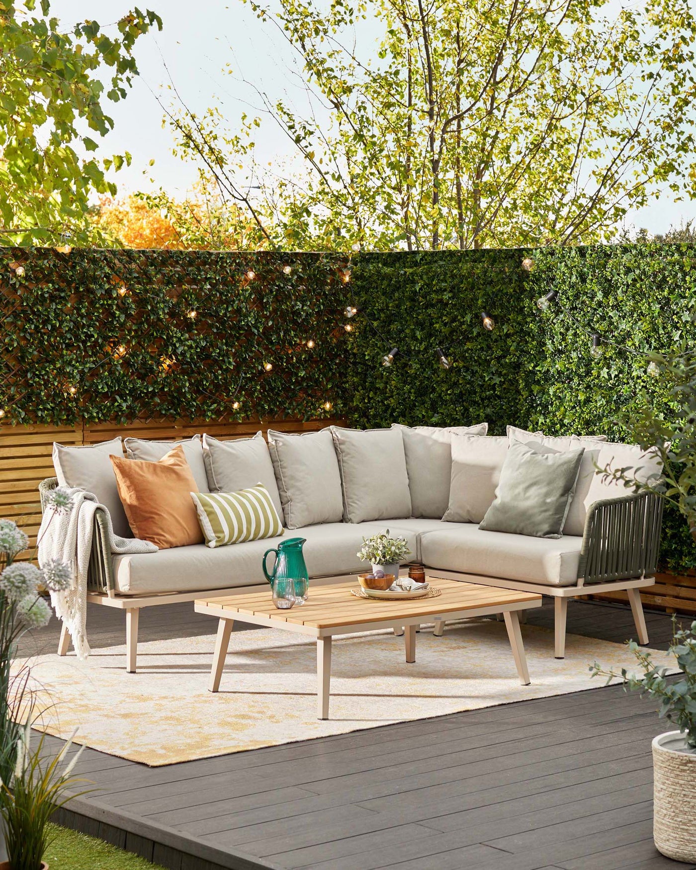 Contemporary outdoor furniture set on a wooden deck, featuring a beige corner sectional sofa with plush seat and back cushions, paired with a modern rectangular wooden coffee table. Decorative accents include assorted throw pillows in earth tones, a fringed throw blanket, a blue glass pitcher, and small potted plants. A cream and yellow patterned area rug lies beneath the table, enhancing the cosy ambiance.