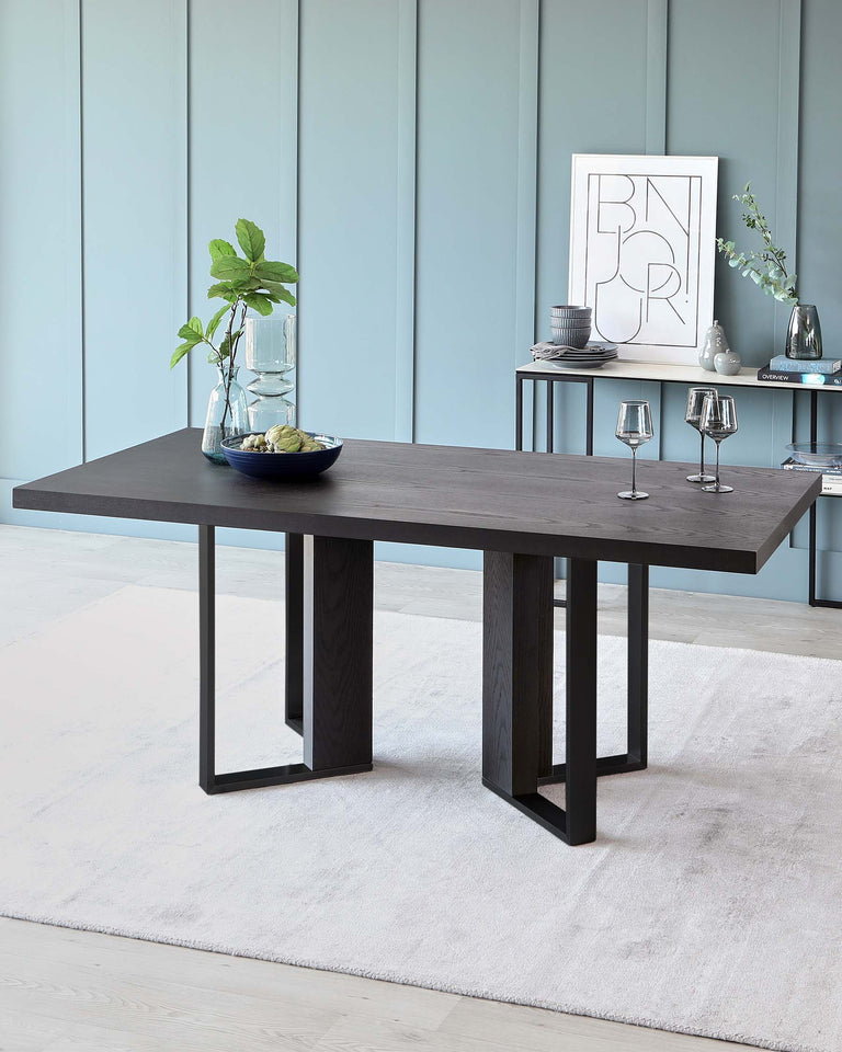 A modern dark wooden dining table with a simple rectangular top and four sturdy block legs, set on a light-coloured rug, styled with decorative items including a clear vase with green foliage, and two elegant stemmed wine glasses.