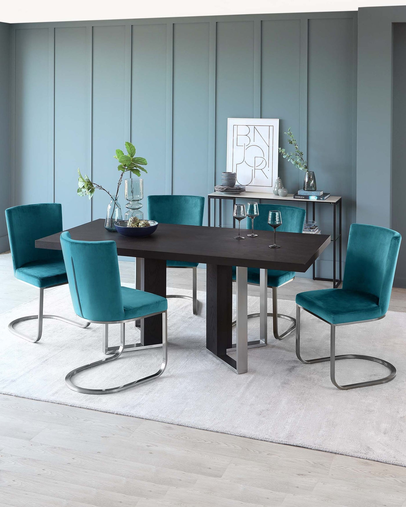 A modern dining set featuring a dark wood rectangular table with chunky legs paired with six teal velvet upholstered chairs with sleek curved metal bases. A matching dark wood console table with decor items and framed artwork is placed against a panelled wall in a muted blue hue. The setup is completed with a light grey area rug and hardwood flooring.