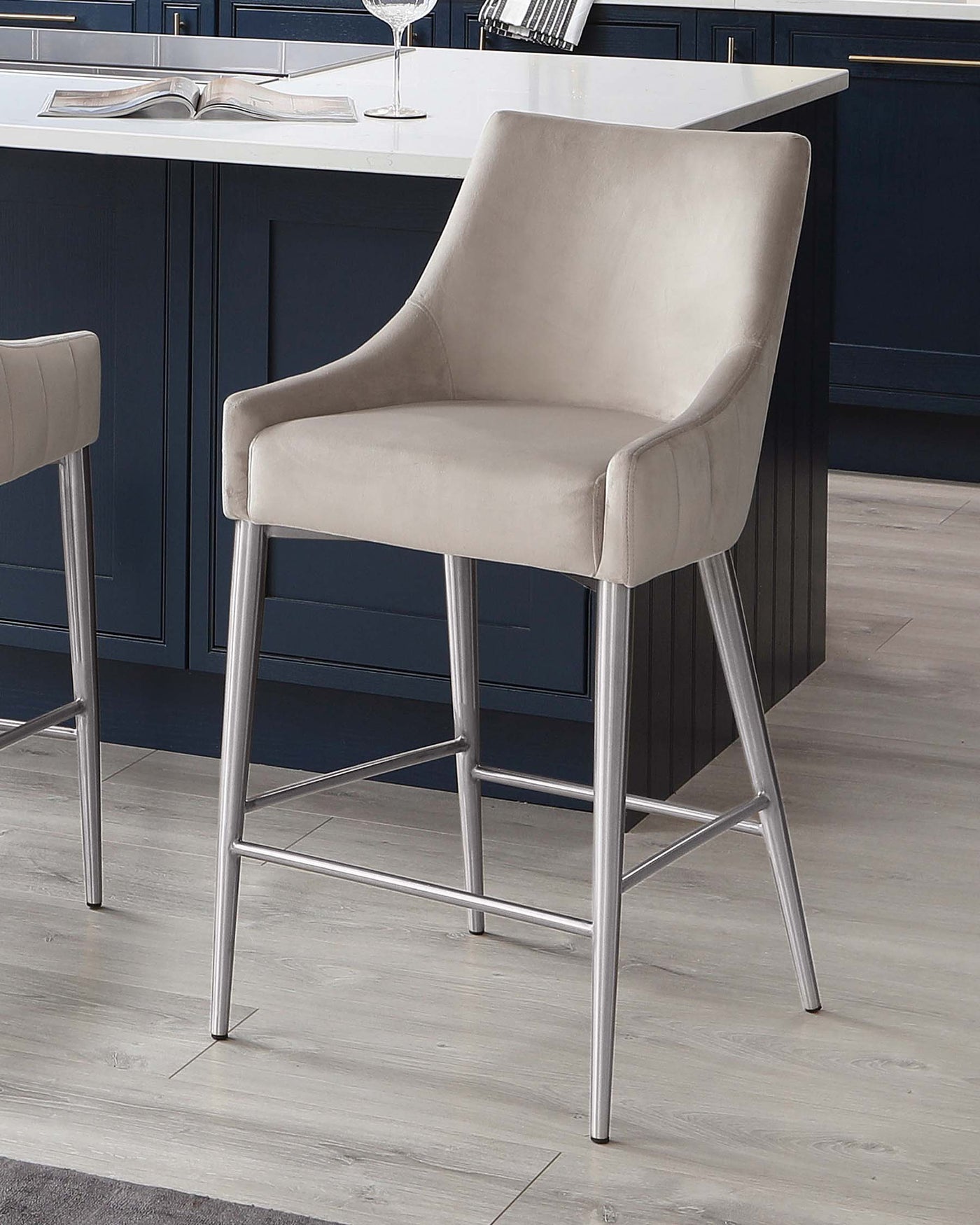 Elegant modern barstool with beige upholstery and sleek chrome legs, featuring a comfortable backrest and armrests. Positioned next to a kitchen island with dark blue cabinetry and white countertop.