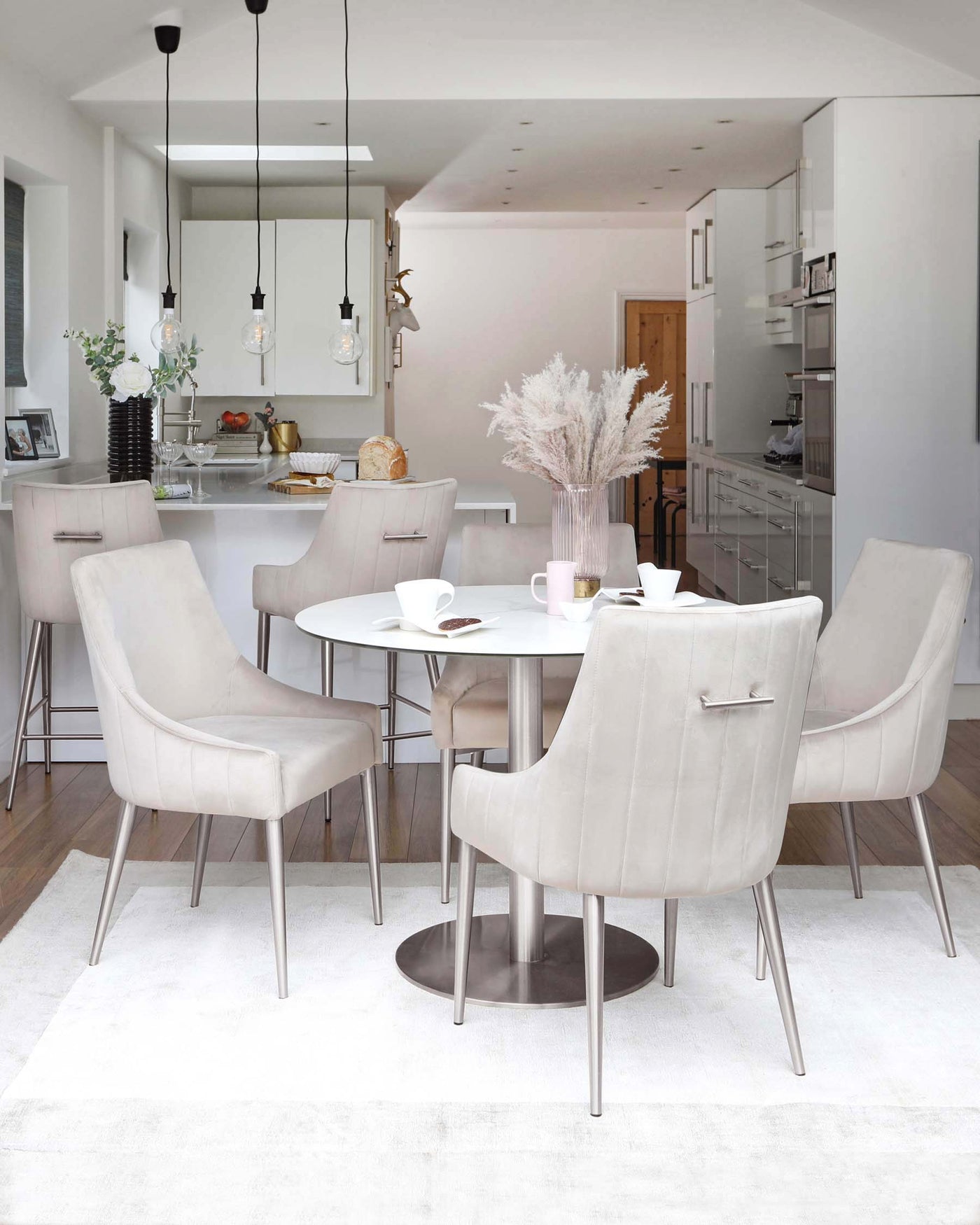 The image features a contemporary dining set consisting of a round, white table with a polished metal base, surrounded by four plush, velvet upholstered chairs in a light beige hue with sleek metal legs. The set is arranged on a soft, textured off-white area rug, complementing the modern and minimalist aesthetic of the space.