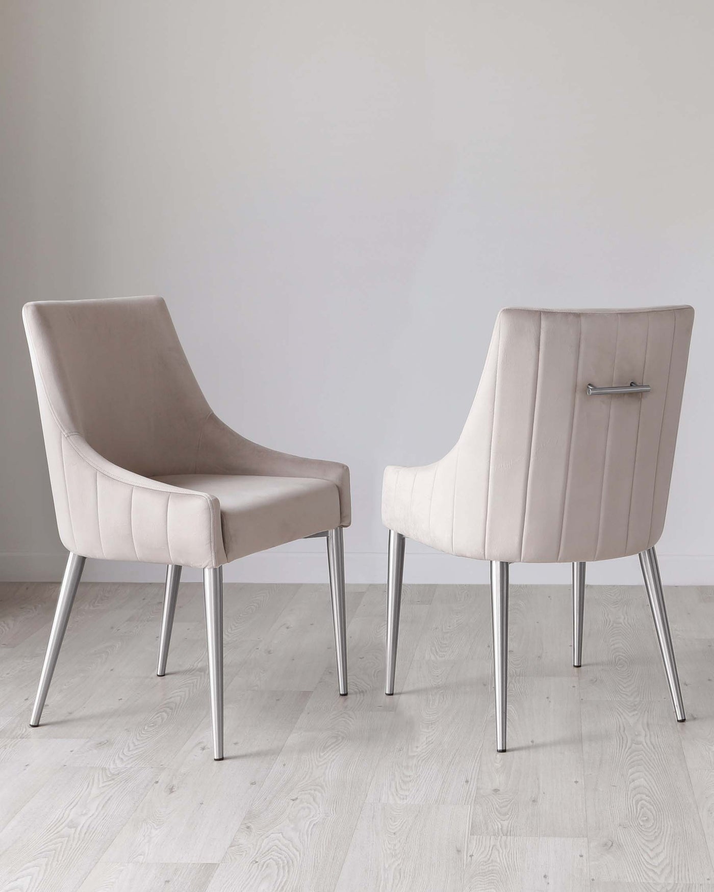 Two modern upholstered dining chairs with sleek, silver metal legs, featuring a gently curved backrest and comfortable seat cushion in an elegant light taupe fabric. The design includes subtle horizontal stitching on the backrest, adding a touch of sophistication. Chairs are positioned on a light hardwood floor against a plain white background.