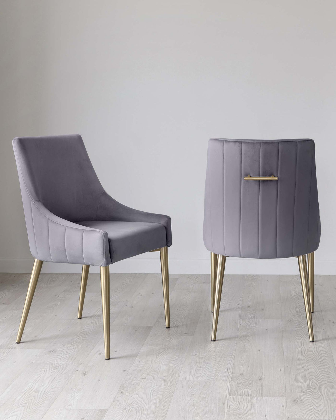 Two elegant modern dining chairs with a luxurious grey fabric upholstery and tapered gold metal legs. The chair on the left features a high back with a slight wingback design and no armrests, while the chair on the right has a lower back with vertical stitching details and an accent handle on the top. Both chairs exude a contemporary sophistication suitable for a chic dining room setting.