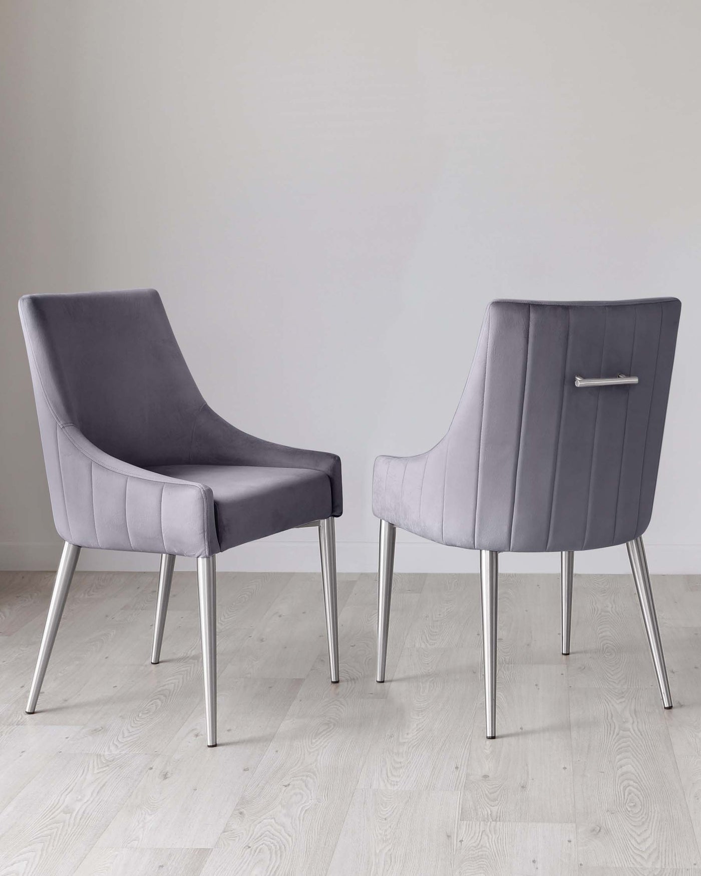 Two modern dining chairs with sleek grey upholstery and horizontal channel tufting. The chairs have slightly curved backrests and are supported by polished chrome legs. The design presents a blend of comfort and contemporary elegance, displayed on a light hardwood floor against a white wall background.