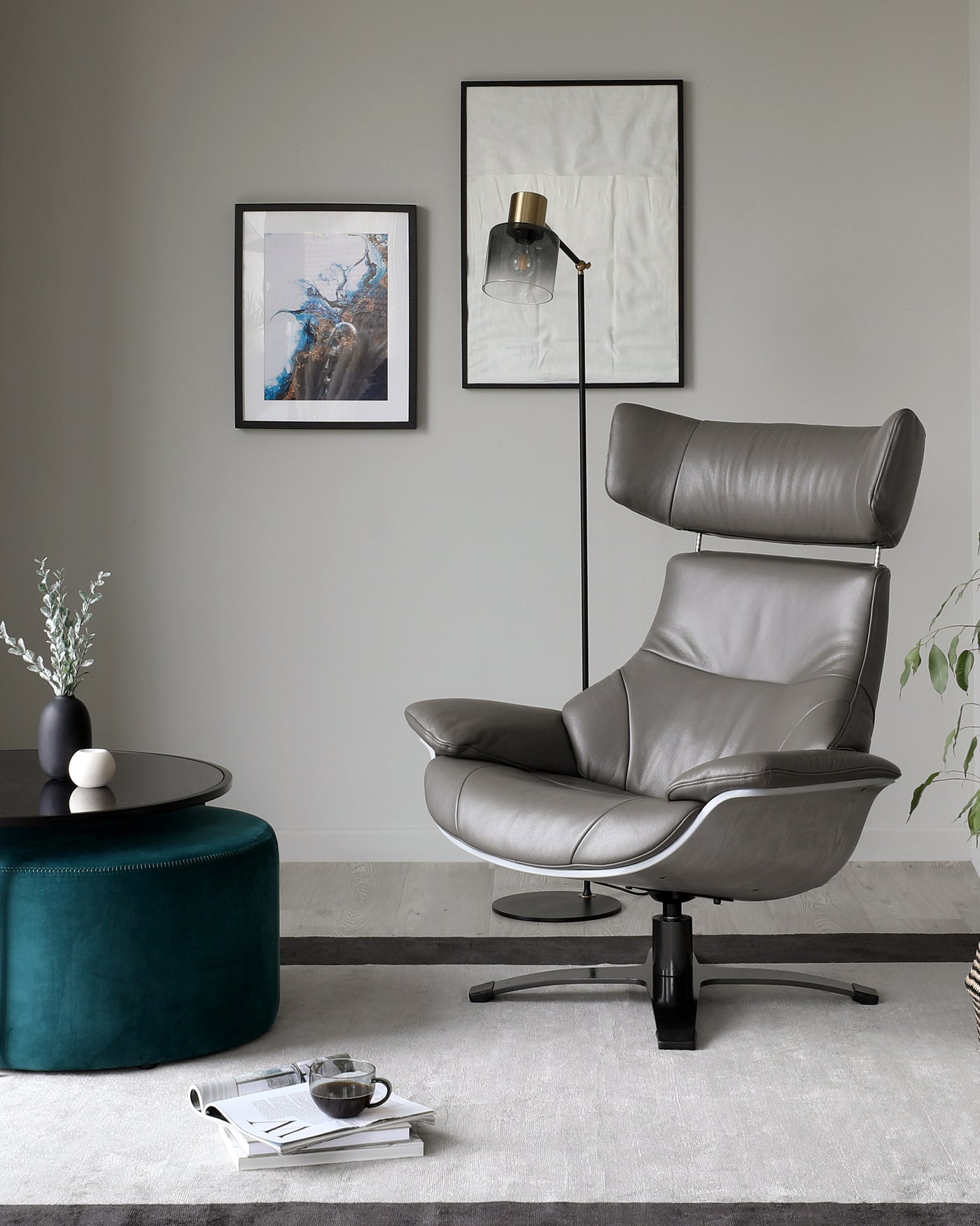 A modern, minimalist room featuring a luxurious grey leather recliner chair with a wide seat, high back, and sleek metal base. In front of it is a round, black coffee table on a textured area rug. To the side is a vibrant teal pouffe, and a tall floor lamp with a bell-shaped glass shade and brass accents casts warm light into the space.
