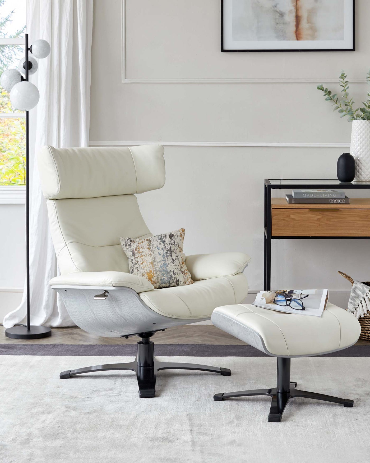 Modern cream leather reclining chair with a matching footstool, featuring a curved wooden base and black metal details. A sleek black and wood side table sits next to it, adorned with decorative items and books.
